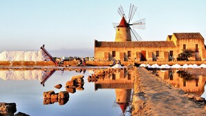 Un moulin à vent, un bâtiment doté d’un toit en tuiles et un grand tas de matériau blanc près d’une étendue d’eau calme.