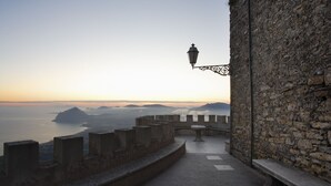 Un mur de pierres surmonté d’une lanterne, un chemin pavé et une vue sur la mer et les montagnes lointaines.