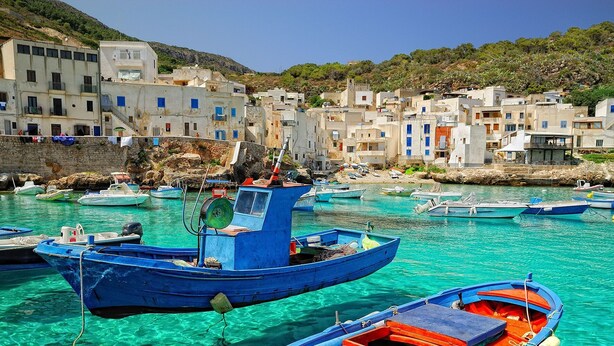 Un village côtier où des bateaux sont amarrés dans des eaux turquoise et limpides, entouré de bâtiments blancs traditionnels rehaussés d’éléments bleus.