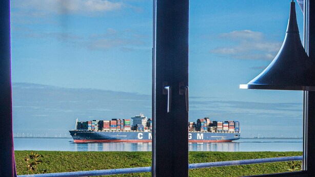 Vista de un buque de carga con el nombre "CMA CGM" en su lateral, atracado en un puerto con cielo despejado y una zona verde al frente.
