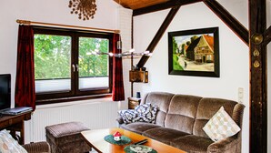 Una habitación acogedora con techo de madera, un ventanal con cortinas rojas, un sofá con cojines estampados, una mesita con un control remoto y un cuadro enmarcado de una casa en la pared.