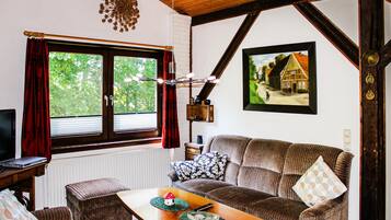 Ein gemütlicher Raum mit Holzbalkendecke, einem großen Fenster mit roten Vorhängen, einer Couch mit gemusterten Kissen, einem kleinen Tisch mit Fernbedienung und einem gerahmten Bild von einem Haus an der Wand.