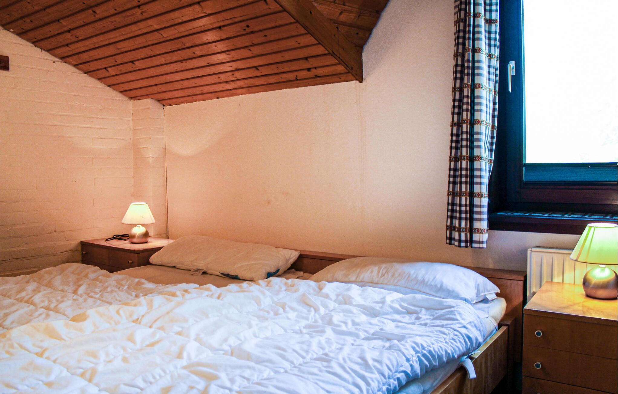 Una camera da letto con soffitto in legno, una finestra con tende a quadri, una lampada da comodino e un letto con biancheria bianca.