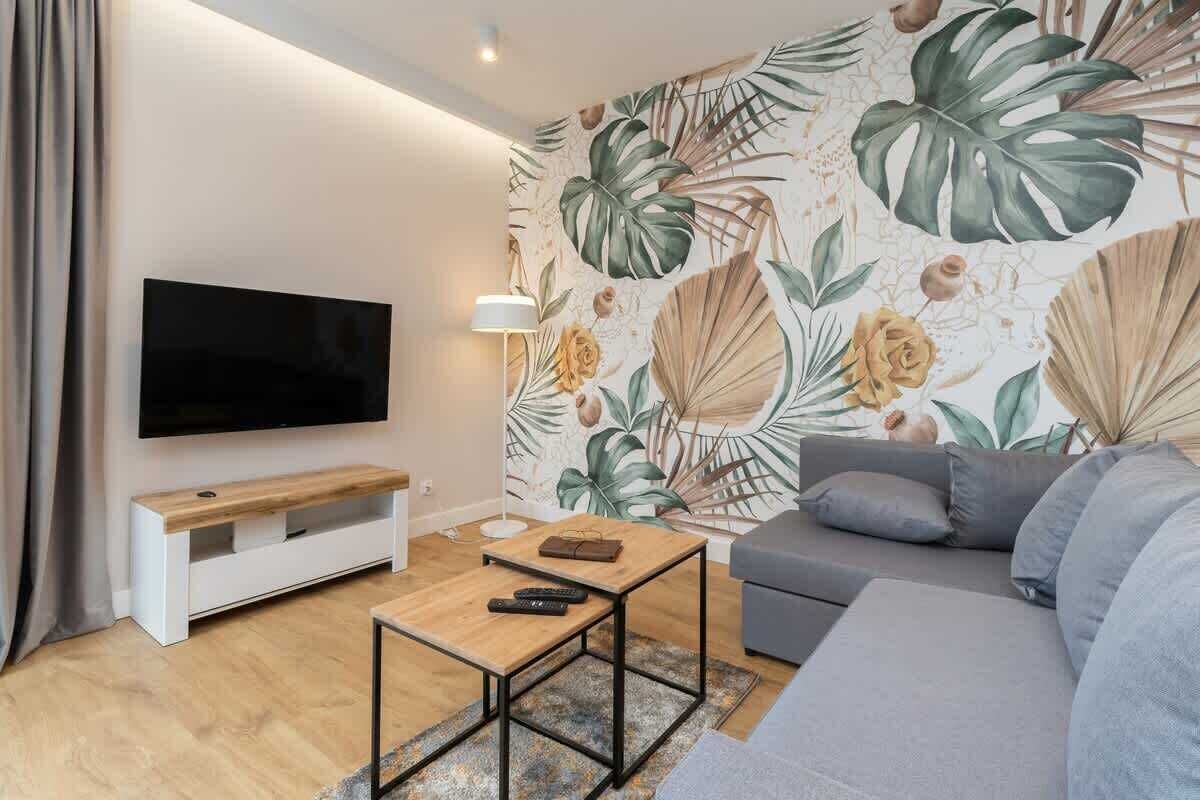 Un salón moderno con un televisor de pantalla plana, una mesa de centro de madera, un sofá gris y un mural con hojas y flores tropicales.