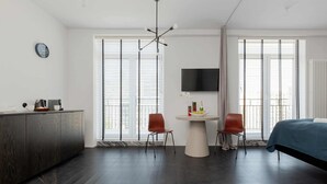 Une chambre d’hôtel moderne avec un plancher en bois foncé, une table ronde avec deux chaises, un lit recouvert d’une courtepointe bleue, un téléviseur fixé au mur et de grandes fenêtres équipées de stores.