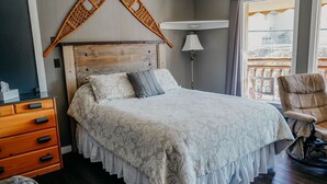 A bedroom with a wooden headboard, a bed with patterned bedding, a wooden dresser, a wheelchair, and a balcony view.