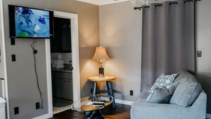 A modern living room with a flat-screen TV mounted on the wall, a glass coffee table, a grey sofa, and a lamp on a small table.