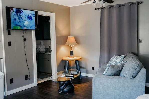 A modern living room with a flat-screen TV mounted on the wall, a glass coffee table, a grey sofa, and a lamp on a small table.