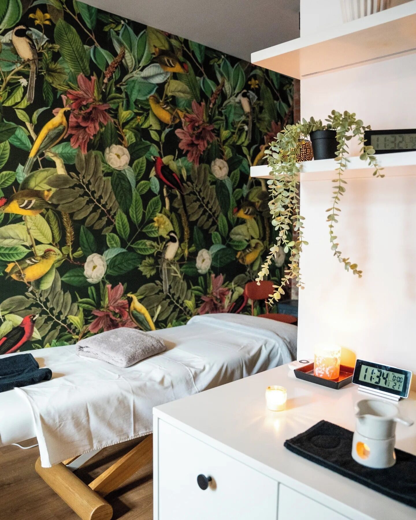 A bedroom with a mural of birds and foliage, a bed, a bedside table with a clock, and a small shelf with a plant.
