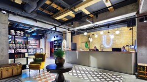 Una hall con pavimento a scacchi, una reception, una poltrona gialla e una libreria con libri e oggetti decorativi.
