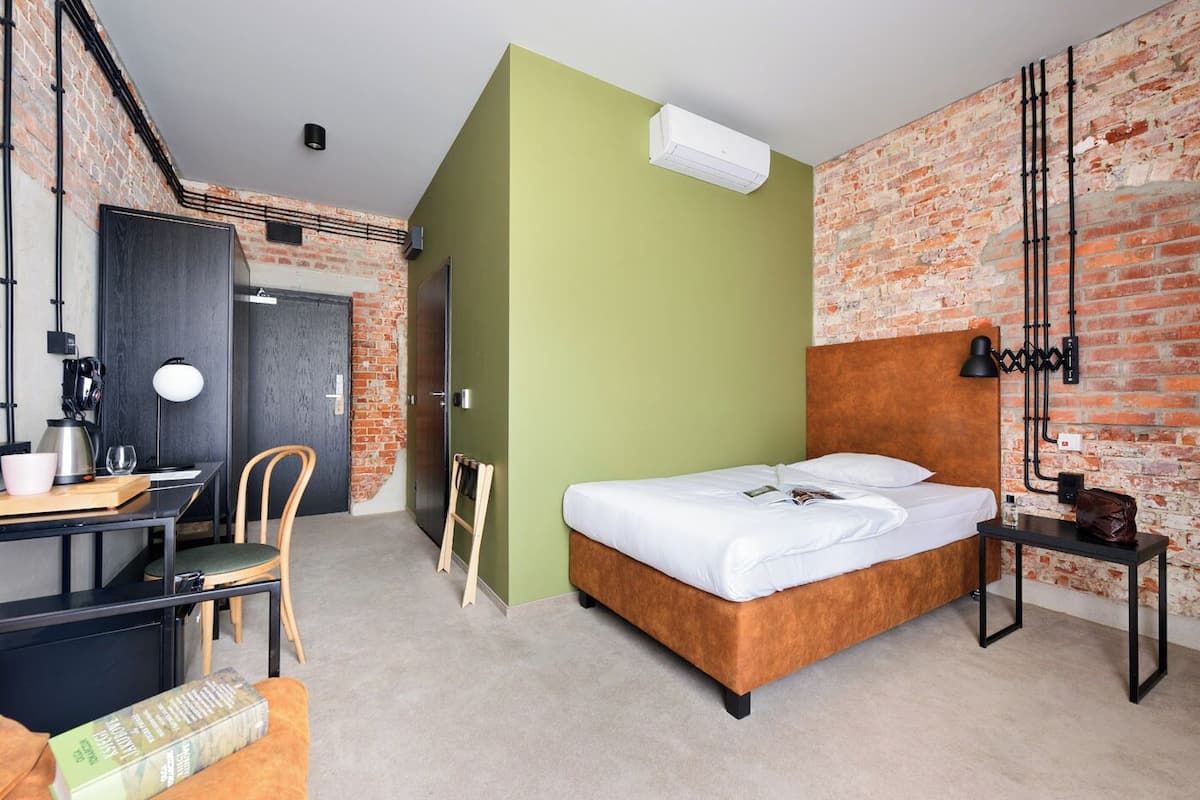 Quarto de hotel moderno com parede de tijolos, cama com roupa de cama branca, escrivaninha com cadeira e uma pequena mesa com abajur.