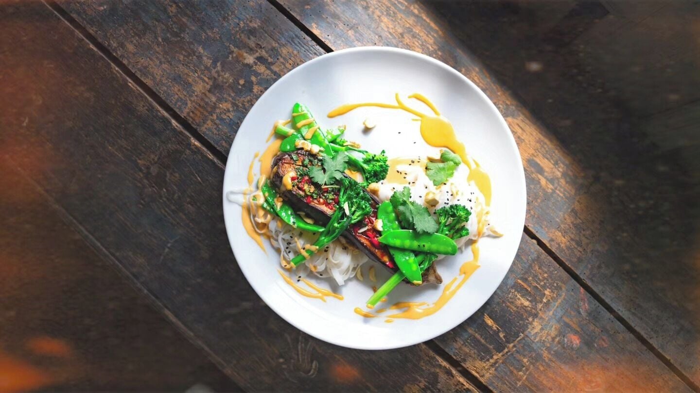 A white plate with a dish of food, including greens and a piece of meat, on a wooden table.
