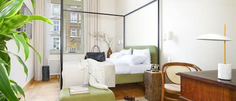 Una cama con dosel, con cabecera tapizada en verde, una mesita de noche de madera, una silla de mimbre y un escritorio con lámpara.