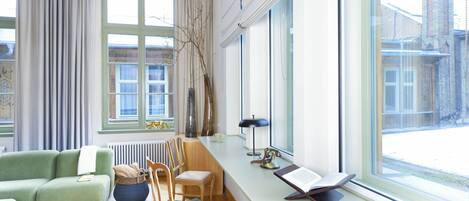 Una sala de estar moderna con un sofá verde, una silla de madera, una mesita con una lámpara y un ventanal con vistas a un edificio.