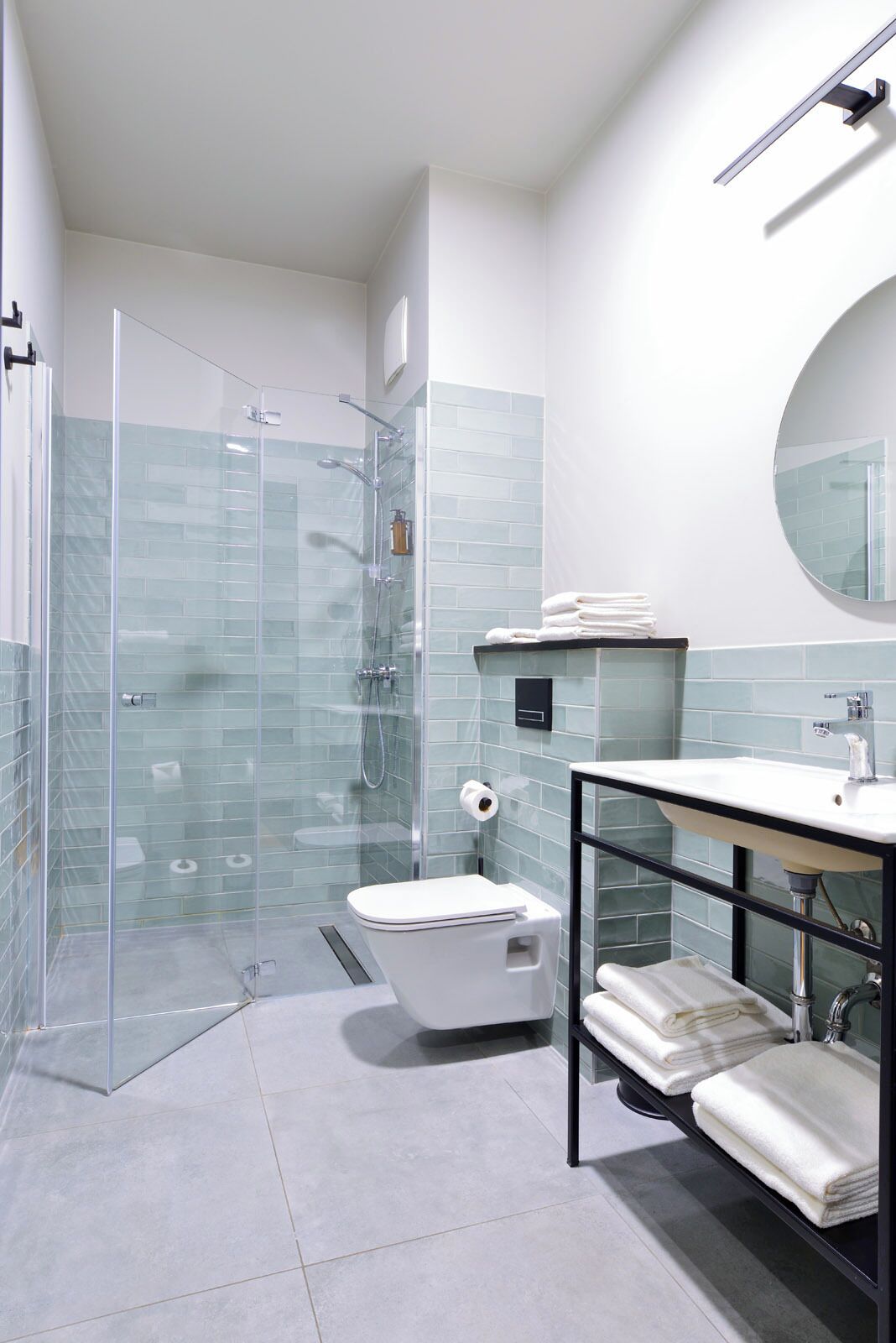 Un baño moderno con ducha envuelta en vidrio, inodoro blanco, lavamanos con estructura negra y espejo redondo.