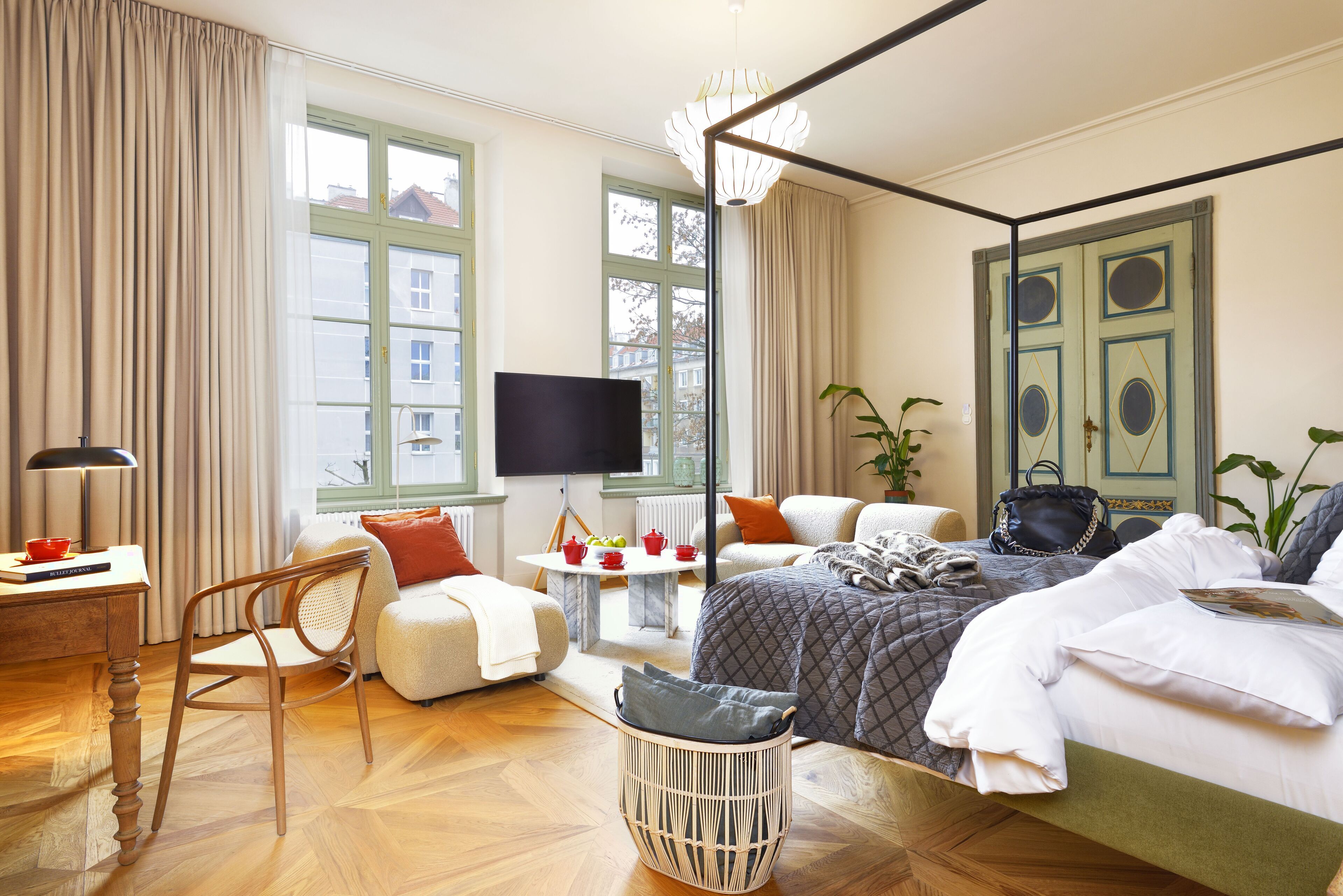 Habitación de hotel bien iluminada con una cama grande, televisión, mesa de comedor con vasos rojos, silla y una cesta de mimbre.