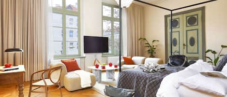 A well-lit hotel room with a large bed, a TV, a dining table with red cups, a chair, and a wicker basket.