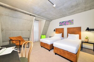 A hotel room with two beds, a desk, a chair, a small table with a lamp, and a painting on the wall.
