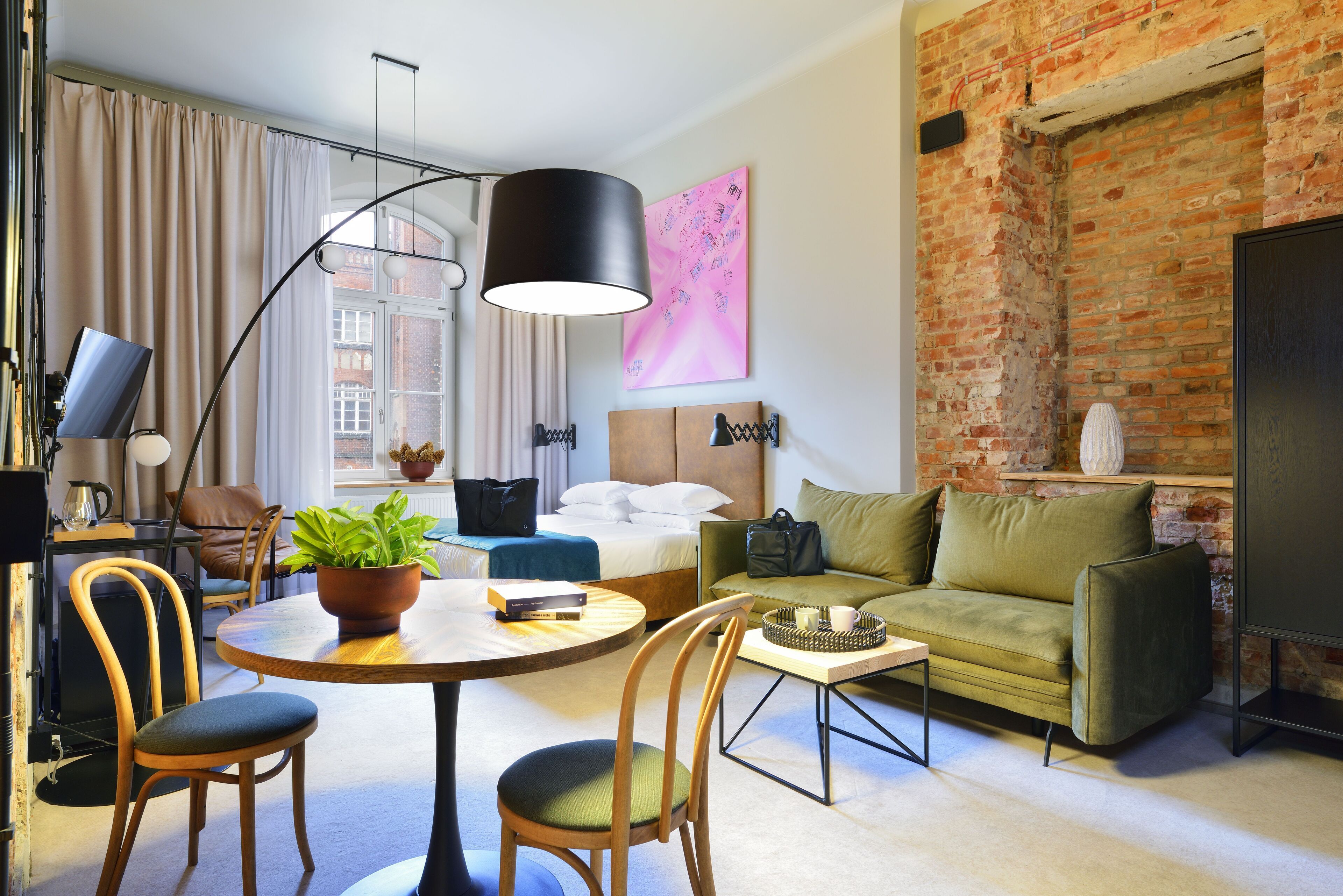 Une chambre d’hôtel moderne avec un mur en briques, un lit recouvert de draps blancs, un canapé vert, une table à manger ronde avec des chaises et une grande fenêtre avec des rideaux.