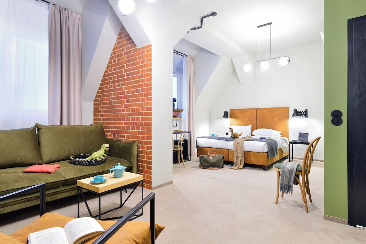 Quarto de hotel moderno com design estilo loft, apresentando um sofá verde, uma cama com cabeceira de madeira e uma pequena área de trabalho.