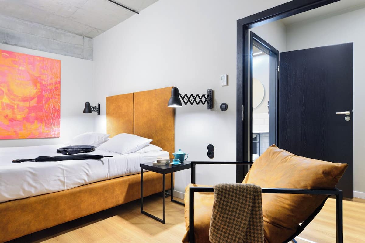 Quarto de hotel moderno com cama de madeira, uma mesa pequena com luminária, uma cadeira xadrez e um quadro abstrato grande.