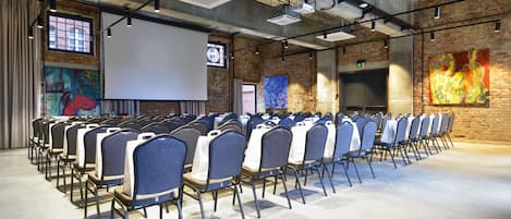 Konferenssihuone, jossa on riveissä tuoleja, projektorikangas ja tiiliseinä, jossa on abstraktia taidetta.