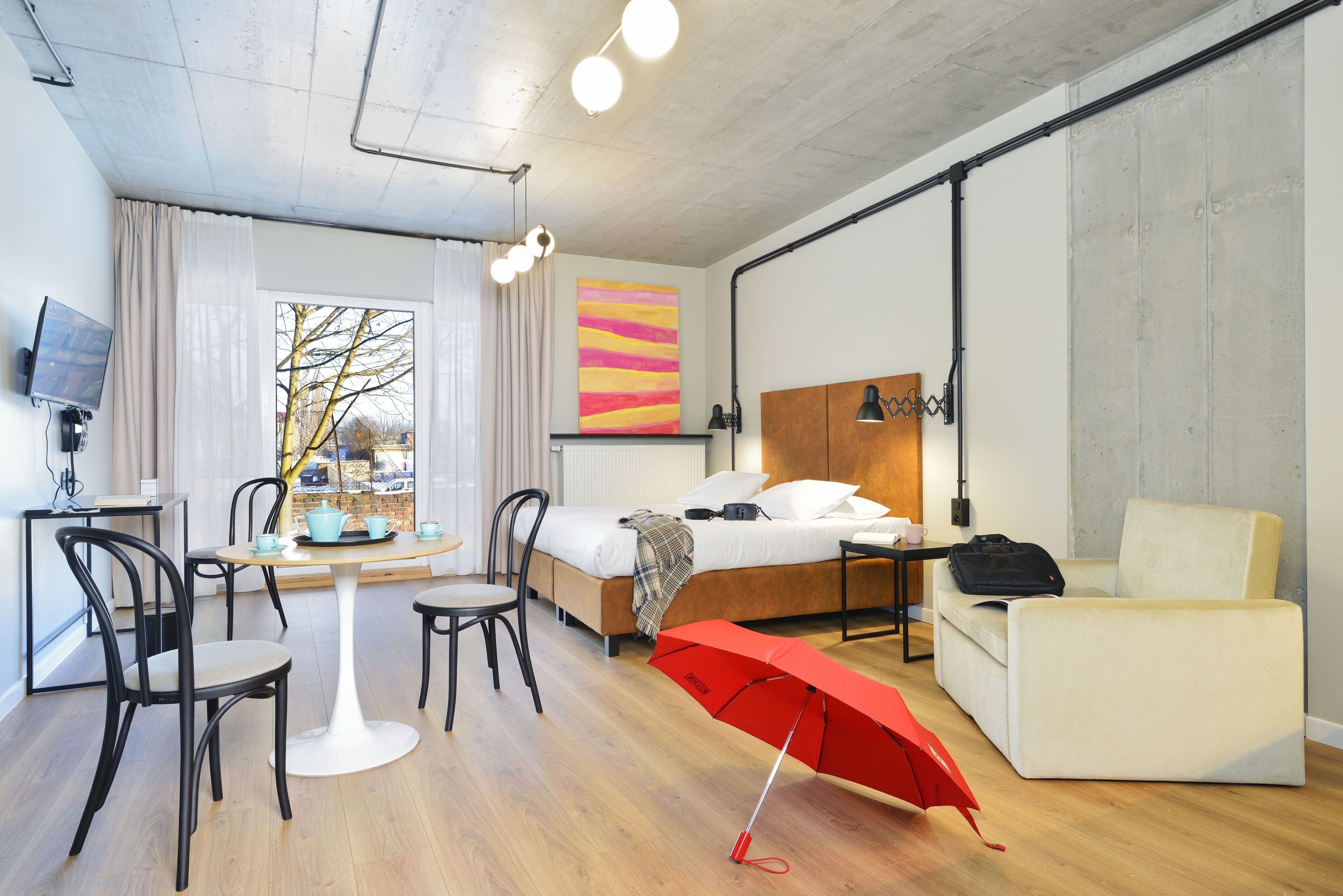 Una habitación de hotel moderna con cama, escritorio, mesa de comedor con sillas, un paraguas rojo y un ventanal con vistas a árboles.