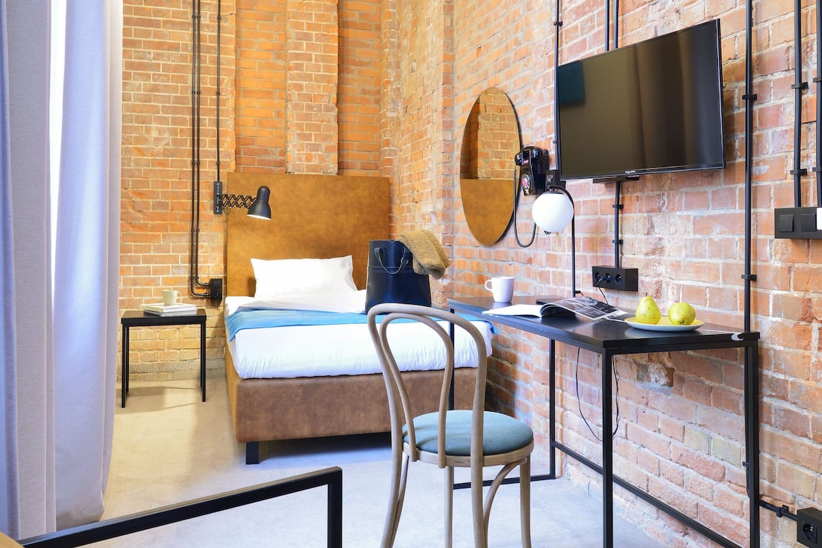 Quarto de hotel moderno com paredes de tijolo aparente, uma cama grande, uma escrivaninha com cadeira, televisão e espelho.