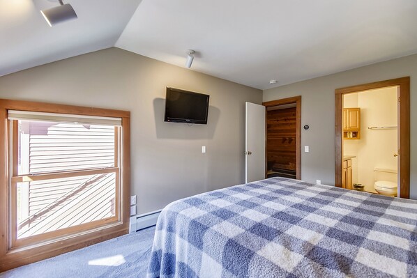 A bedroom with a checkered bedspread, a mounted TV, a window with blinds, and a door leading to a bathroom.