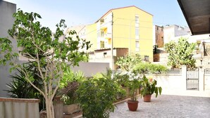 A courtyard with potted plants, a yellow building in the background, and a stone floor. - Casa Vacanze le Palme Netine (Noto)