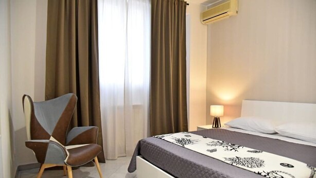 A hotel room with a bed, a chair, a nightstand with a lamp, and an air conditioning unit. - Casa Vacanze le Palme Netine (Noto)