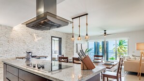 A modern kitchen with a central island, stainless steel range hood, hanging pendant lights, and a view of the ocean through large windows. - Casa Shell, Where the Caribbean Is Part of the View (Playa del Carmen)