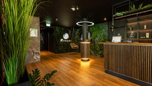 A reception area with a wooden floor, a reception desk with a computer, a hanging chair, and a wall with green foliage. - Rioca Munich Posto 3 (Munich)