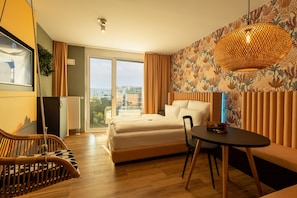 A modern hotel room with a large bed, a dining table, a wicker chair, and a view of the city.