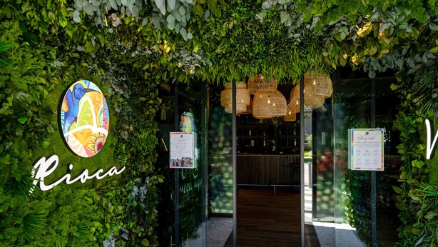 A restaurant entrance with a green leafy archway, a colorful logo, and a menu sign. - Rioca Munich Posto 3 (Munich)