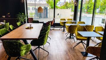 A modern dining area with wooden tables and chairs, large windows offering a view of the street, and hanging pendant lights.
