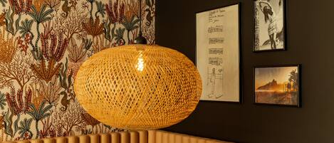 Une pièce avec un papier peint à motifs, un luminaire en fibres tressées et des œuvres d’art encadrées accrochées au mur.