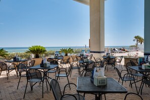 An outdoor seating area with tables and chairs, overlooking a beach and the sea.