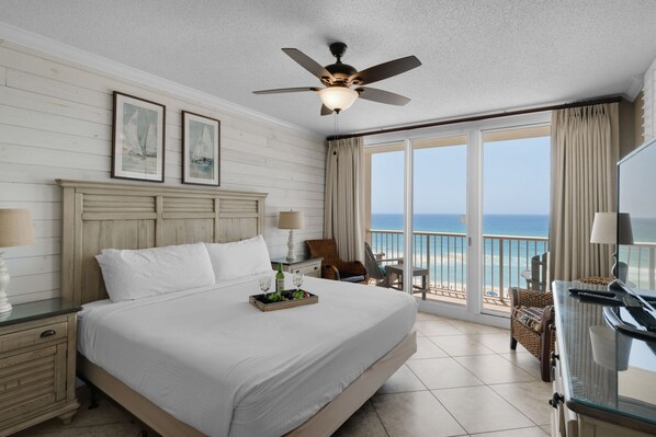 A hotel room with a large bed, a ceiling fan, a view of the ocean, and a balcony with wicker furniture.