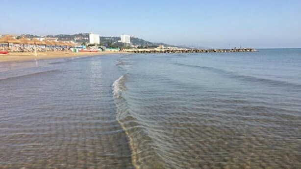 A beach with clear shallow waters, a pier extending into the sea, and buildings in the background. - Casolare Fonte di Moro (Abruzzo)
