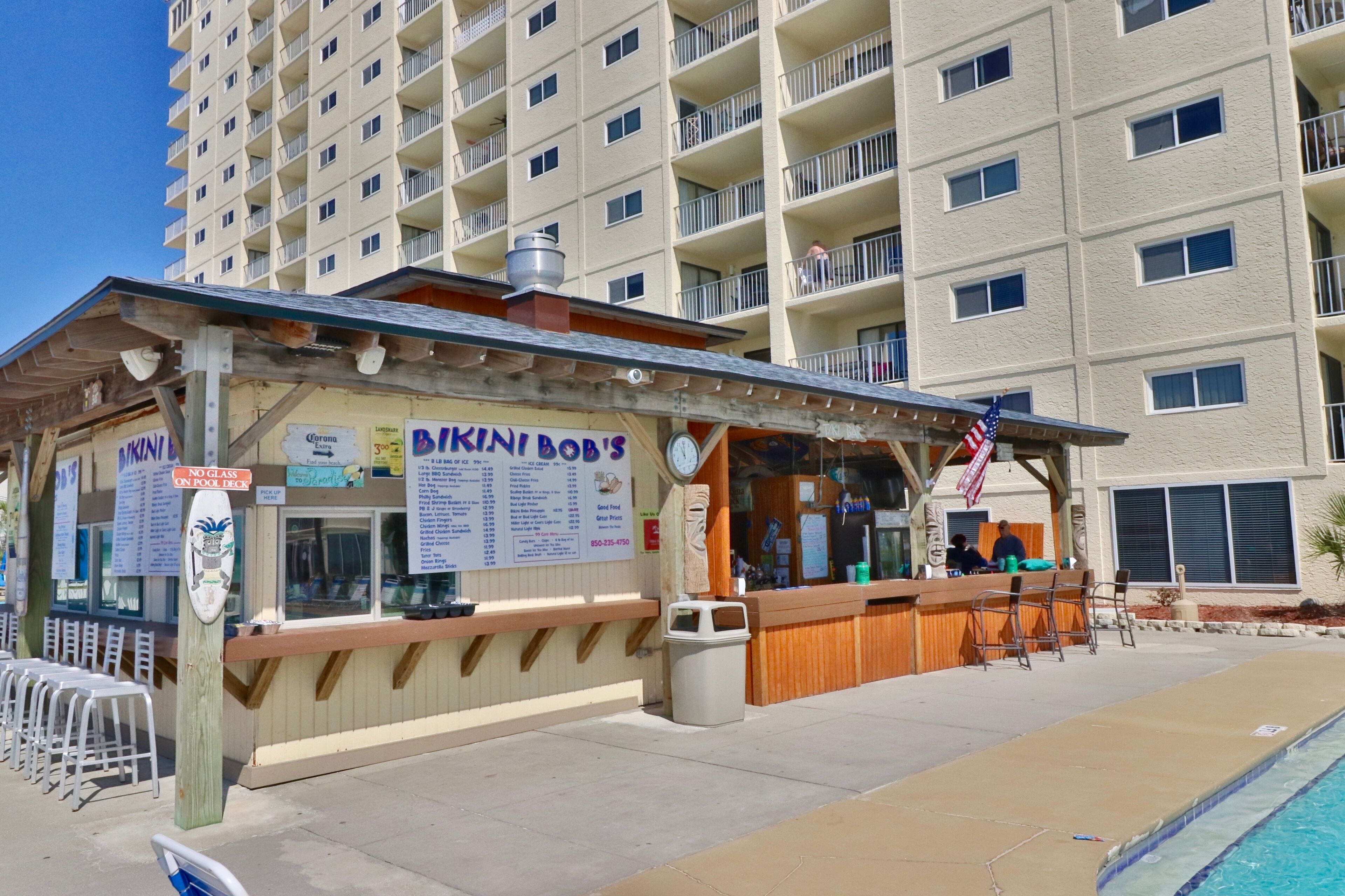 Un bar situé en bord de mer, appelé « Bakin' Bob's », avec un espace salon près de la piscine et une vue sur un grand immeuble d'habitation en arrière-plan.