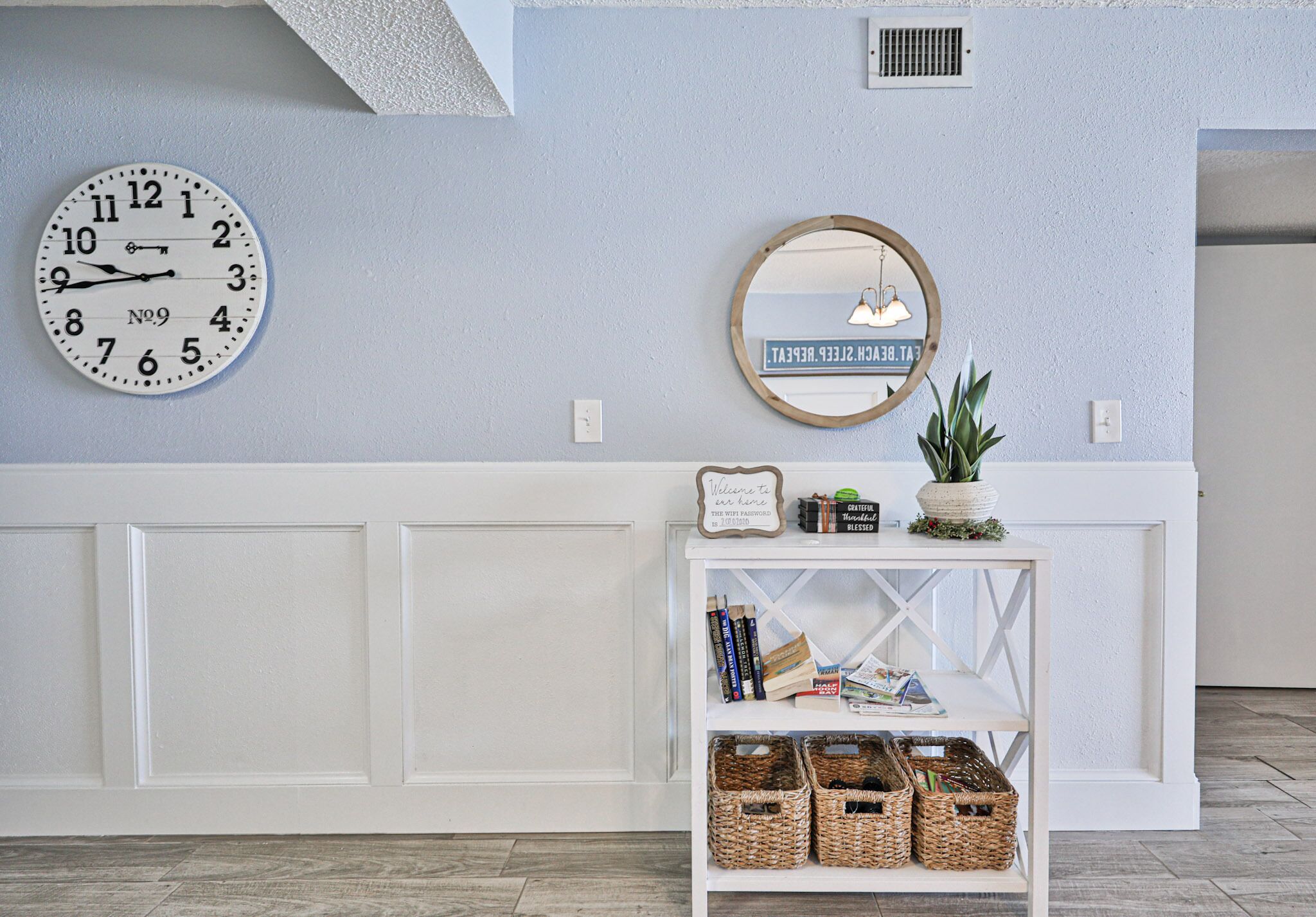 A room with a white console table, a round mirror, a clock, and wicker baskets.