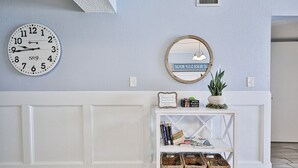 Una habitación con una mesa consola blanca, un espejo redondo, un reloj y cestas de mimbre.