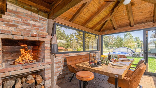 Eine Außenküche mit Kamin aus Ziegelsteinen, einem Holztisch und Stühlen, mit Blick auf einen Garten mit Pool und Bäumen.