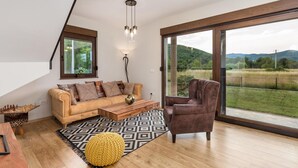 Ein modernes Wohnzimmer mit Sofa, Sessel, Couchtisch aus Holz und einem großen Fenster mit Blick auf eine Wiese und Hügel.