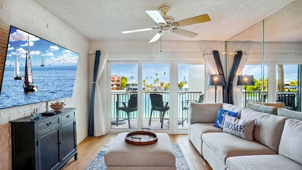 A living room with a large TV, a ceiling fan, a sofa, and a balcony with outdoor furniture.
