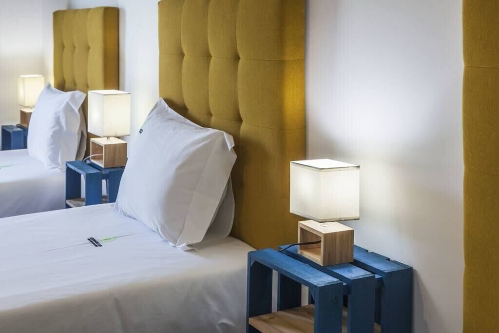 A hotel room with two beds, each with a tufted headboard, bedside tables with lamps, and a bedside table with a smartphone.