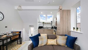 A modern apartment with a dining area, a sofa with 'FAMILY' pillows, a kitchen, and a view of the sky. - Host & Stay | Captain's Retreat (Whitby)