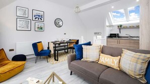 A modern living room with a sofa, armchairs, and a dining table. There are framed pictures on the wall, a clock, and a kitchen area with a window. - Host & Stay | Captain's Retreat (Whitby)
