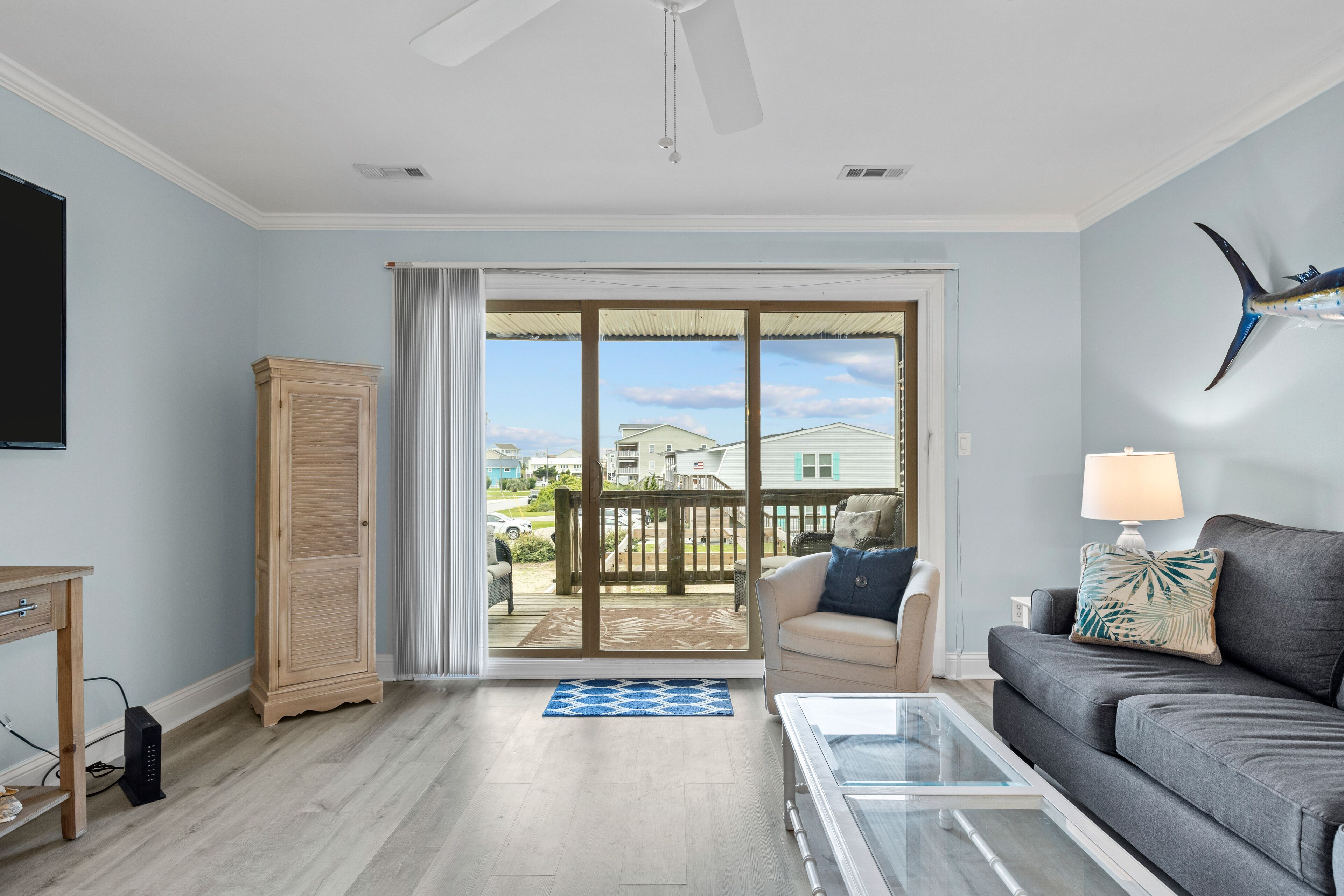 Un salon comprenant un canapé, une table basse, un téléviseur et donnant sur un balcon avec une terrasse en bois et des maisons en arrière-plan.